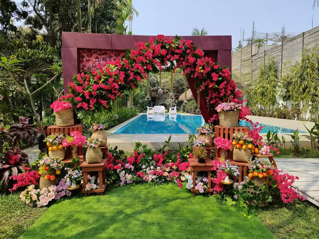 An outdoor wedding ceremony setup in Bangladesh with a large red floral arch, a swimming pool in the background, and chairs set up for a couple.