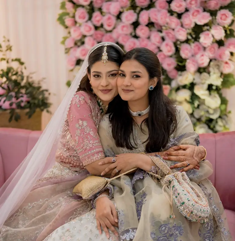 Bride in a blush pink embellished outfit embracing her friend on a pink velvet sofa in front of a floral backdrop at a 300 Feet Purbachal wedding reception venue by Look N Feel - Event Solutions.