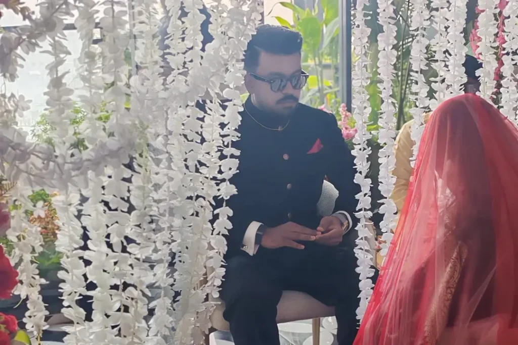 Groom in a black prince suit and sunglasses seated opposite bride draped in a sheer red veil during a Nikah ceremony at Skyfall Mohakhali by Look N Feel - Event Solutions.